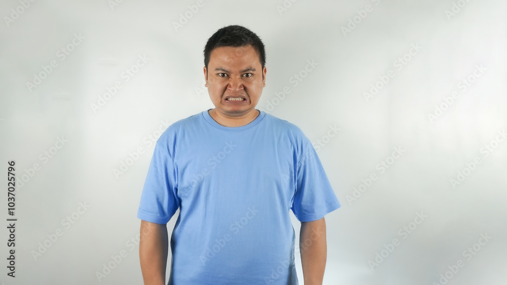 A young Indonesian man wearing a blue t-shirt with an angry face facing forward A young Indonesian man wearing a blue t-shirt with an angry face facing forward