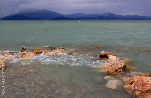 View of the azure water of Lake Garda with stones in the foreground and mountains in the background against a stormy sky in Sirmione, Italy