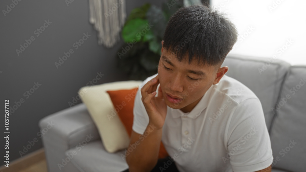 Handsome young chinese man sitting indoors in a modern living room wearing a white shirt looking contemplative with his hand on his chin Handsome young chinese man sitting indoors in a modern living room wearing a white shirt looking contemplative with his hand on his chin