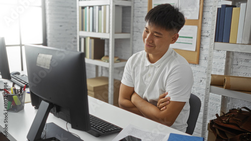 Young man in a white shirt sitting with arms crossed in a home office, looking at a computer screen, surrounded by books and shelves, conveying a relaxed yet focused mood