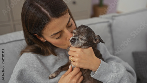 Young hispanic woman affectionately kissing her senior chihuahua in a cozy living room.
