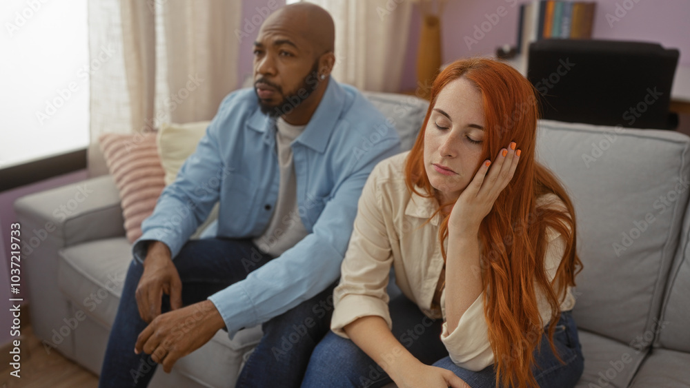 Obraz premium Interracial couple sitting together indoors in a living room, woman with hand on face, both looking thoughtful