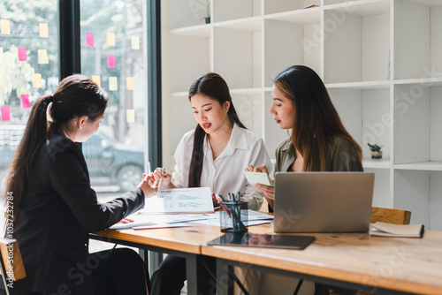 Collaborative business meeting with three women discussing project details at modern office table. They are engaged in conversation, showcasing teamwork and professionalism