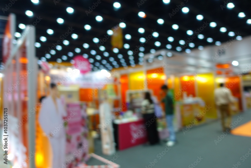 Naklejka premium Abstract blurred image of people visiting a large education exhibition hall or trade show. The scene features vibrant booths and visitors walking along the aisles under bright ceiling lights.