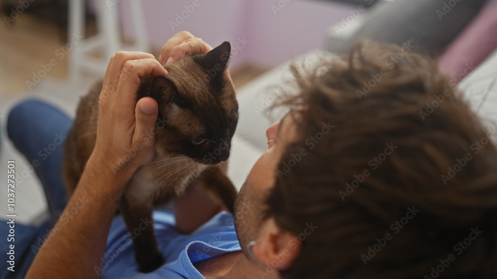 A young hispanic man affectionately pets a siamese cat in a cozy indoor room, exemplifying a heartwarming human-animal bond. A young hispanic man affectionately pets a siamese cat in a cozy indoor room, exemplifying a heartwarming human-animal bond.