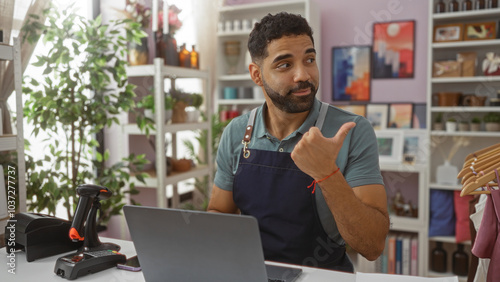 Young hispanic man in apron working at home decor store pointing towards shelves with plants and art in background