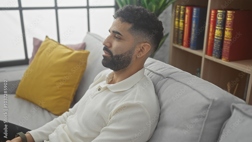 A young hispanic man with a beard wearing a casual shirt sits thoughtfully in a modern living room. A young hispanic man with a beard wearing a casual shirt sits thoughtfully in a modern living room.
