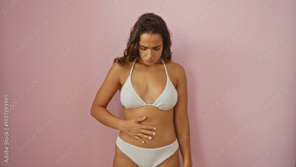 Young brunette woman in white bikini over isolated pink background looking down at her stomach with a concerned expression Young brunette woman in white bikini over isolated pink background looking down at her stomach with a concerned expression