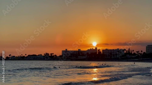 Sunset over the sea in Ayia Napa, Cyprus, with gentle waves and people swimming. In the distance, a city silhouette adds peaceful autumn scene during the travel velvet season.