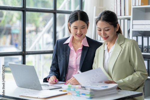 Collaborative Success: Two smiling Asian businesswomen review documents together in a modern office, showcasing teamwork and professional collaboration.