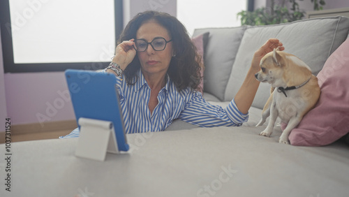 A middle-aged woman with glasses focused on a tablet while petting a chihuahua on a couch indoors. photo