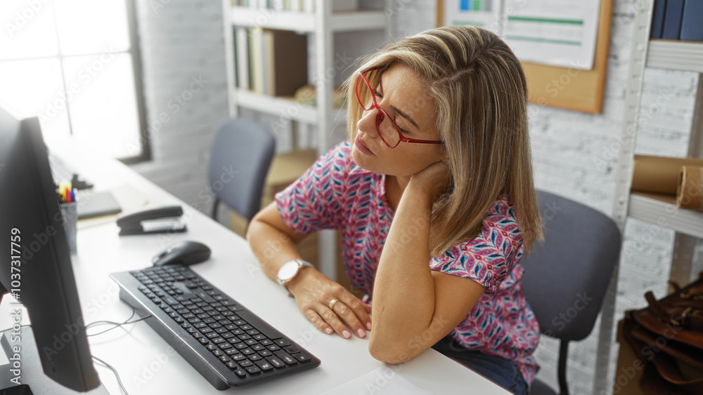 Woman working at computer in office, wearing glasses, feeling neck pain, seated at desk with keyboard and mouse, indoor setting with shelf and documents in the background Woman working at computer in office, wearing glasses, feeling neck pain, seated at desk with keyboard and mouse, indoor setting with shelf and documents in the background
