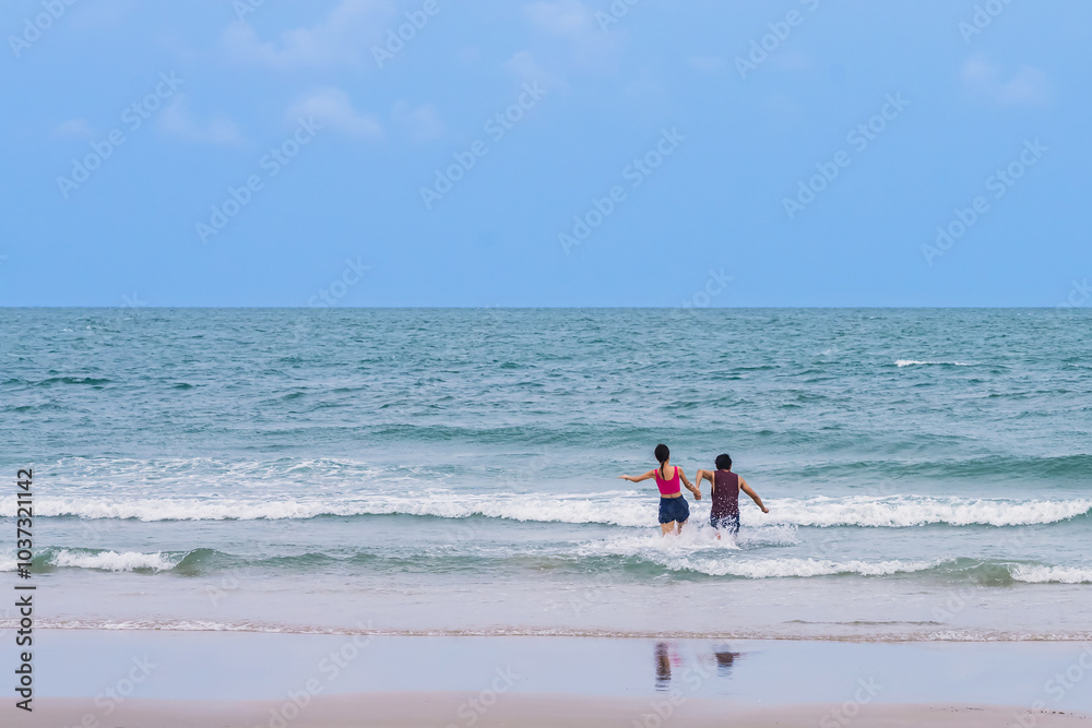 Back view of happy carefree couple holding hands and running in water at beach. Couple plays in ocean waves. Happy young couple playing in sea water. Guy and girl splashing together, beach vacation. Back view of happy carefree couple holding hands and running in water at beach. Couple plays in ocean waves. Happy young couple playing in sea water. Guy and girl splashing together, beach vacation.