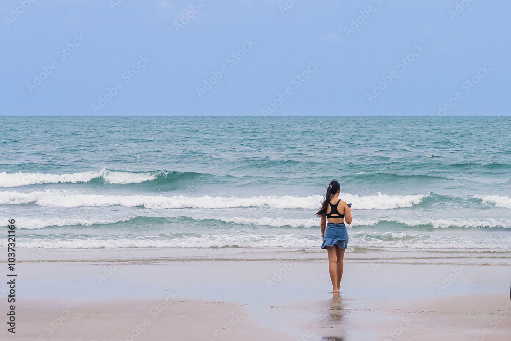 Back view of happy carefree woman using mobile phone at beach. Woman using smartphone standing at beach. Beautiful lovely young woman spending time at beach. Concept of travel, tourism and vacation. Back view of happy carefree woman using mobile phone at beach. Woman using smartphone standing at beach. Beautiful lovely young woman spending time at beach. Concept of travel, tourism and vacation.