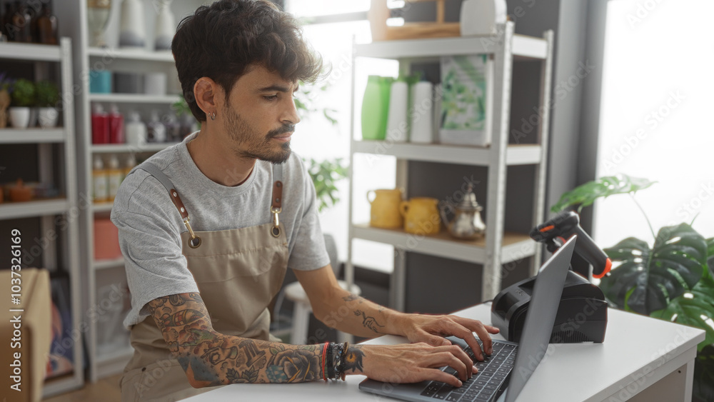 Young hispanic man with beard and tattoos working on laptop in home decor store interior with shelves of decorations Young hispanic man with beard and tattoos working on laptop in home decor store interior with shelves of decorations