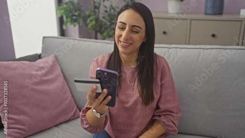 Smiling hispanic woman holding credit card and mobile phone in a cozy home interior.