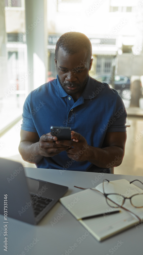 Young african american man in an office setting checking his phone with a laptop and notepad on the desk Young african american man in an office setting checking his phone with a laptop and notepad on the desk