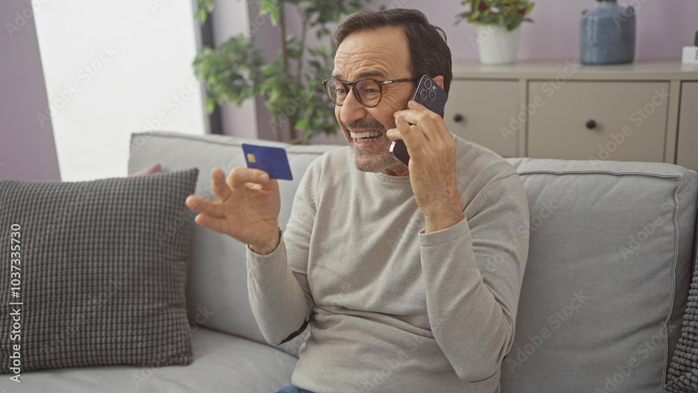 Hispanic man talking on phone holding credit card in a cozy living room Hispanic man talking on phone holding credit card in a cozy living room
