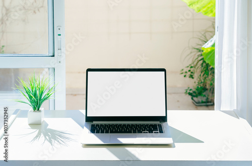 Laptop on a desk with white screen for mockup. There are no people on the scene. Computer next to the window. Stock photographs with clear, bright colors.