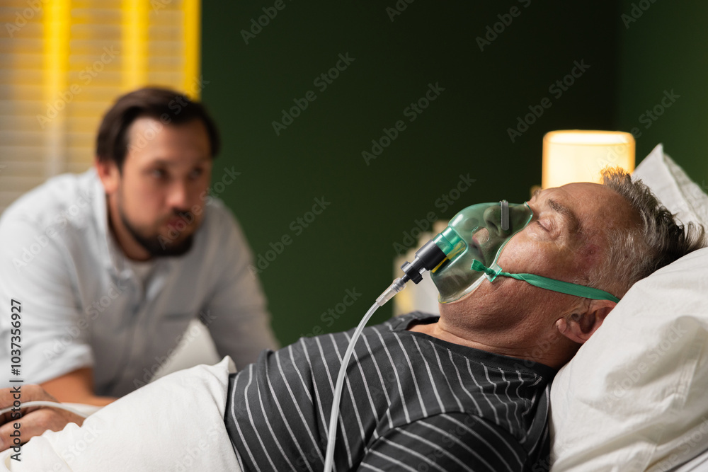 Fototapeta premium A senior man in a hospital bed, using an oxygen mask, is peacefully asleep, his son keeping a watchful eye over him.