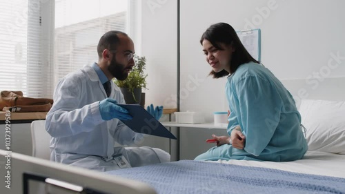Gen Z girl in patient gown sitting on bed in hospital ward and discussing plastic surgery plan on clipboard with male doctor during preoperative consultation