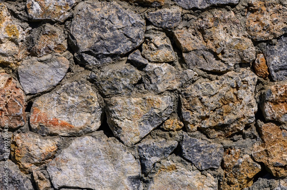 Textured stone wall composed of various sized rocks, showcasing natural colors and patterns under bright daylight Textured stone wall composed of various sized rocks, showcasing natural colors and patterns under bright daylight
