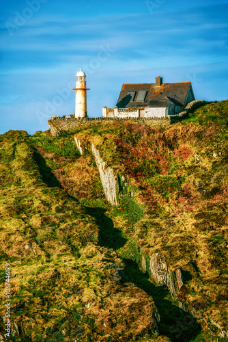 A white lighthouse stands on a rocky cliff, next to a small, white, single-story house with a grey roof. The sky is a clear blue, and the sun is shining brightly. photo