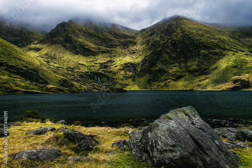 A rocky shore slopes down to a still, dark water lake, with two large, green mountains in the distance. photo