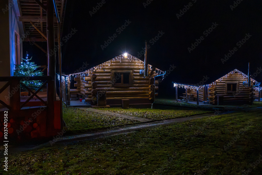 Cozy cabins illuminated by winter lights in a forest clearing at night, a Christmas tree, garland. Cozy cabins illuminated by winter lights in a forest clearing at night, a Christmas tree, garland.