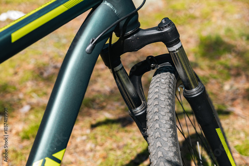 Close-up of a bicycle parked on a dirt trail, with a focus on the kickstand. The bike's rugged tires are well-suited for trail riding, and the scene suggests a forest environment.