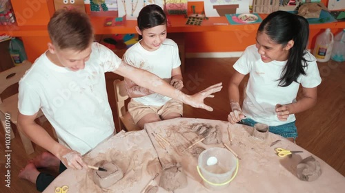 Top view of caucasian boy use color brush put clay on muddy shirt. Diverse high school student playing mudin pottery at table with dough scatter around workshop. Creative activity concept. Edification photo