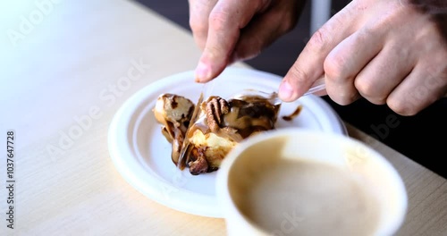 A close up of a woman hands slicing a chocolate caramel cinnamon roll disposable cutlery on a white plate coffee in foreground Captures cozy moment perfect showcasing indulgent desserts cafe vibes.
