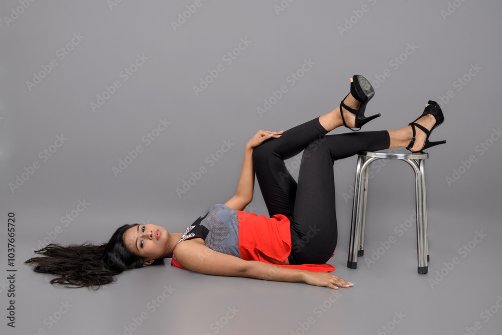 Indian female model in casual wear wearing red and grey designer top, black jeans and black high heel stiletto posing with a stool against grey background. Female model. Fashion Portrait. Indian female model in casual wear wearing red and grey designer top, black jeans and black high heel stiletto posing with a stool against grey background. Female model. Fashion Portrait.