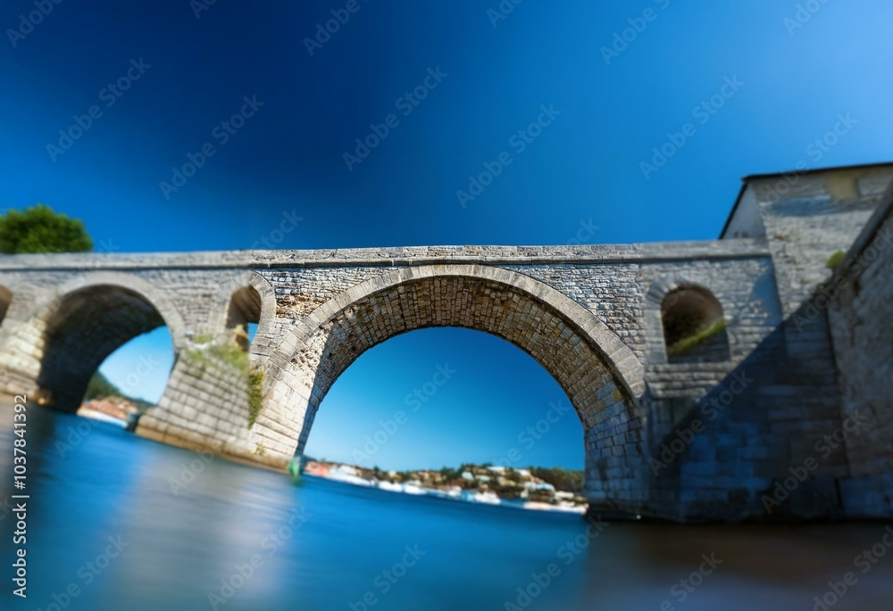 Fototapeta premium An arched stone bridge spans a blue body of water, with a clear blue sky above.