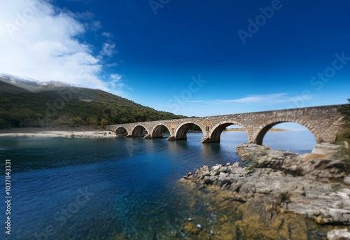 Stone arch bridge spans a calm, clear blue body of water, with a green hillside beyond.