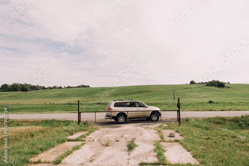 A white SUV is driving steadily down a bumpy dirt road that runs right next to a lush, green grassy field in the beautiful scenery