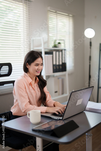 A young woman is sitting at her desk in an office, working on her laptop. She looks focused and engaged, with office documents and a coffee mug beside her, in a bright, modern workspace