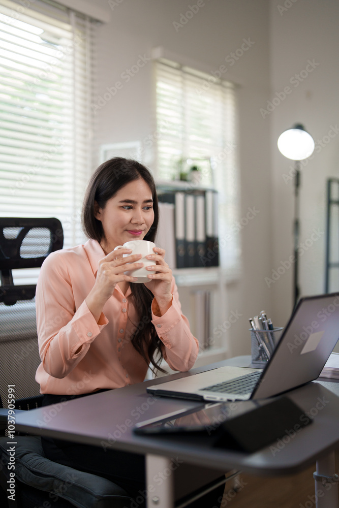 A young woman is sitting at her desk, holding a cup and smiling while looking at her laptop. She is working in a cozy and modern office environment, enjoying a break during her work. A young woman is sitting at her desk, holding a cup and smiling while looking at her laptop. She is working in a cozy and modern office environment, enjoying a break during her work.