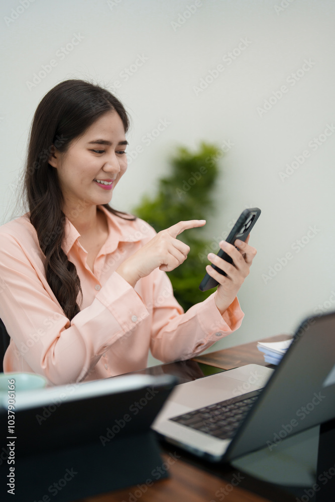 A young woman is sitting at her desk, smiling as she looks at her phone while working on her laptop. She is in a bright and cozy workspace A young woman is sitting at her desk, smiling as she looks at her phone while working on her laptop. She is in a bright and cozy workspace