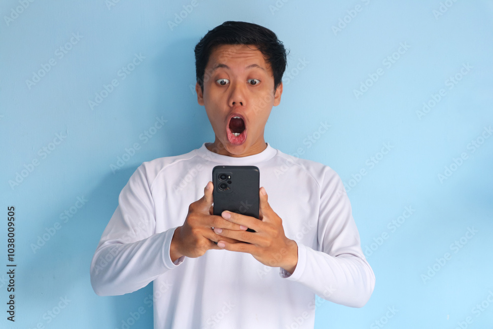 Young Asian man wearing white t-shirt surprised while holding cellphone Young Asian man wearing white t-shirt surprised while holding cellphone