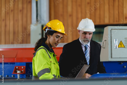 Two people are looking at a laptop, one of them is wearing a yellow jacket. Scene is serious and focused to machine controller board in factory.