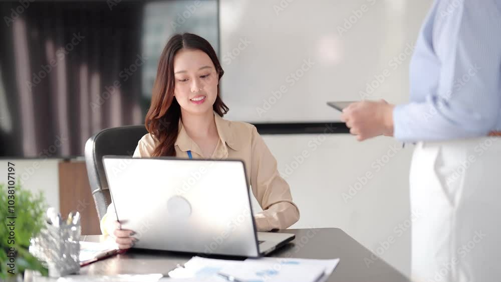Two young women are working together in a coworking space. One woman is sitting and focused on the laptop, discussing or explaining, holding a clipboard Two young women are working together in a coworking space. One woman is sitting and focused on the laptop, discussing or explaining, holding a clipboard