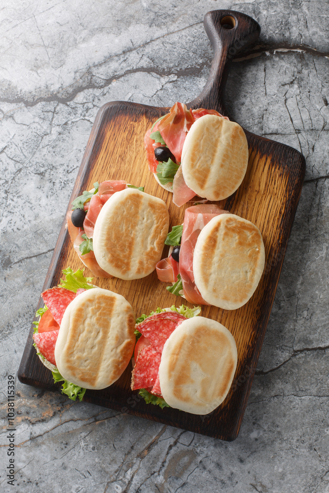 Typical Italian Food Tigella sandwich with salami, ham and fresh vegetables close-up on wooden board on table. Vertical top view from above