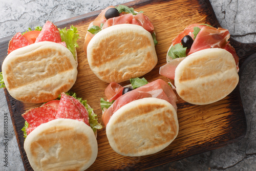 Small italian tigelle sandwiches with sausage, jamon and fresh vegetables close-up on wooden board on table. Horizontal top view from above photo