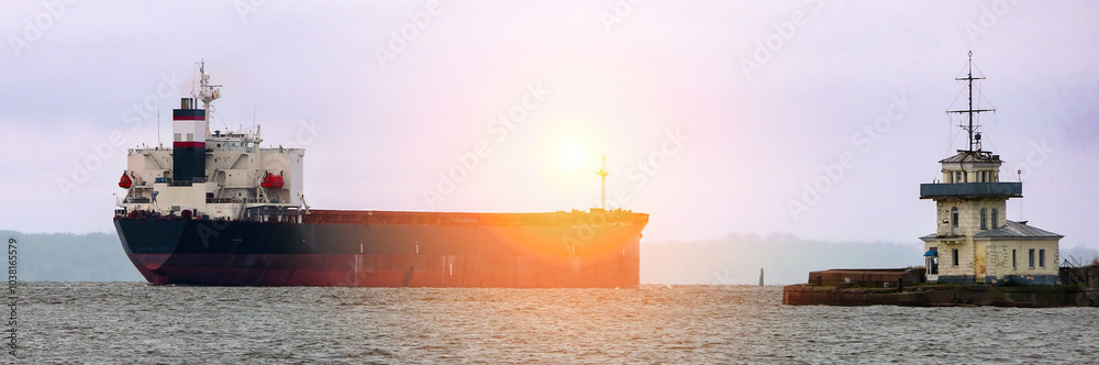 Fototapeta premium A cargo ship navigates through coastal waters near a lighthouse under a twilight sky