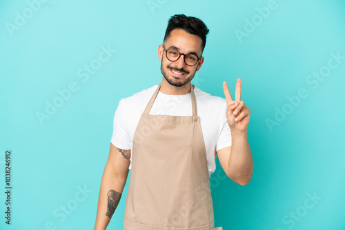 Restaurant waiter caucasian man isolated on blue background smiling and showing victory sign