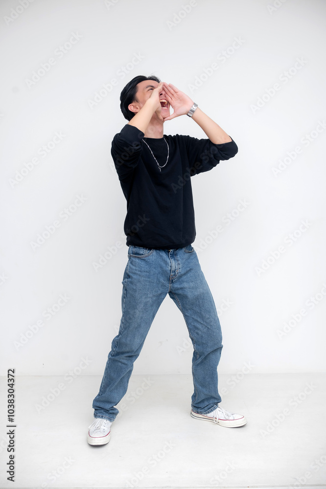Young asian man wearing black shirt and jeans yelling with cupped hands on white background Young asian man wearing black shirt and jeans yelling with cupped hands on white background