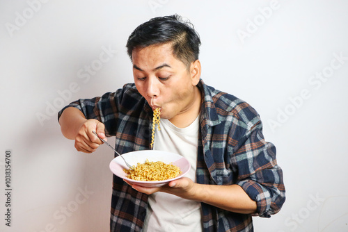 An Asian man in a casual shirt eagerly eats noodles from a plate, his expression filled with delight. The plain white background focuses on his enjoyable dining experience.