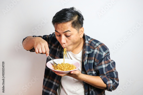 An Asian man in a casual shirt eagerly eats noodles from a plate, his expression filled with delight. The plain white background focuses on his enjoyable dining experience.