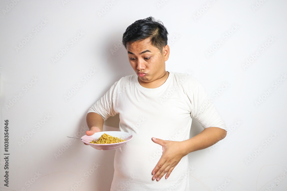 Young Asian man with noodles on plate holding stomach showing belly fat Young Asian man with noodles on plate holding stomach showing belly fat